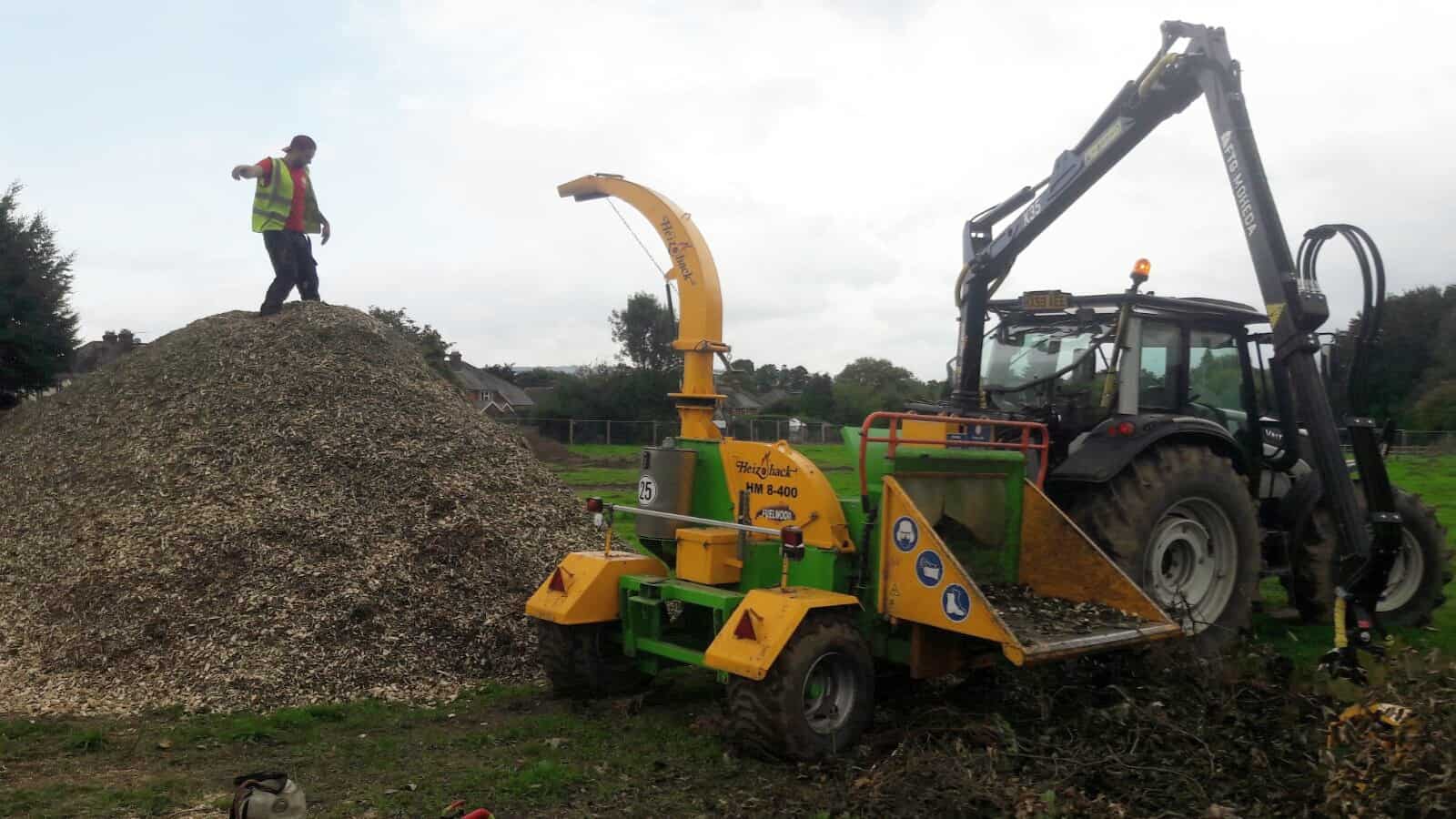 Crane-fed wood chipper with a huge pile of woodchip. A tree surgeon stands on top of the pile.